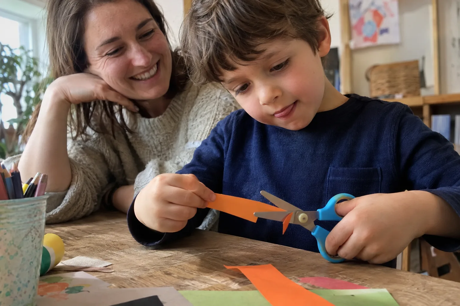 Un garçon découpe une bande de papier coloré avec des ciseaux à bouts ronds tenus dans sa main gauche, encouragé par sa mère.
