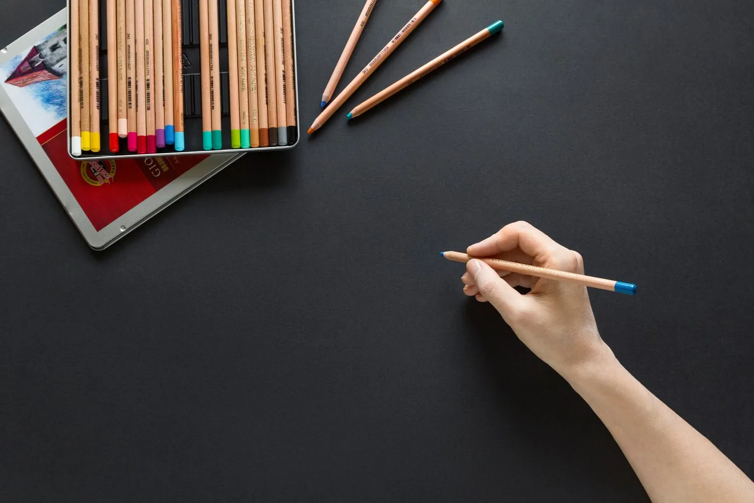 Une main d’adulte tient un crayon de couleur en pince tripode au-dessus d’une feuille noire.