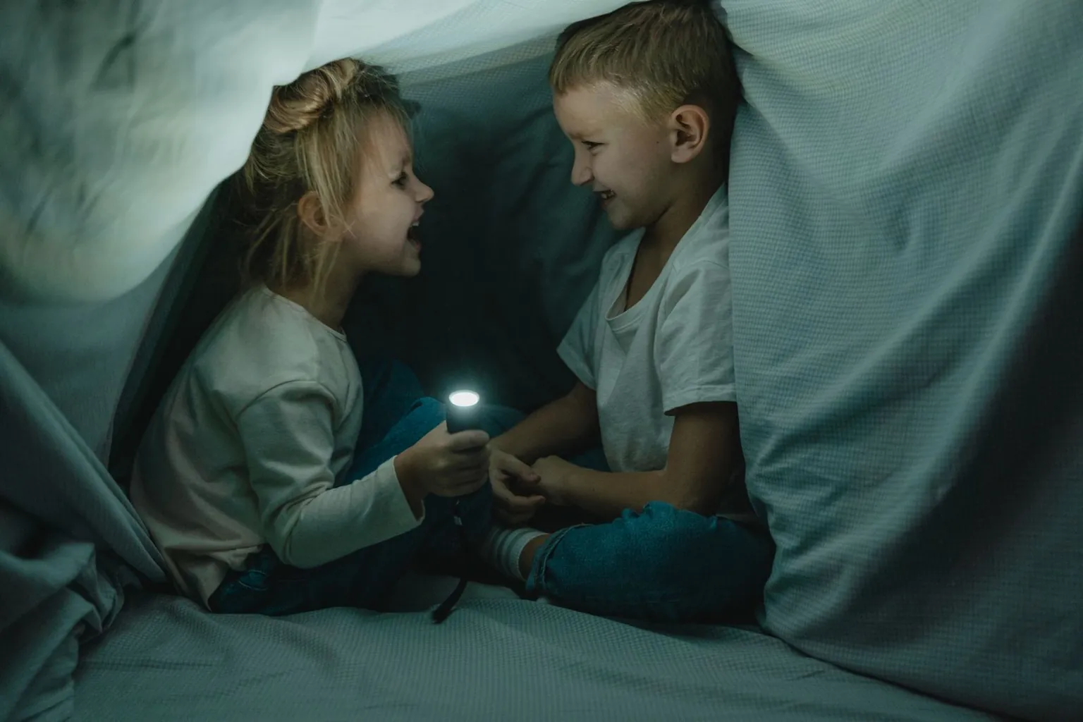 Deux enfants se parlent dans une cabane faite d’un drap, éclairés par une lampe de poche.