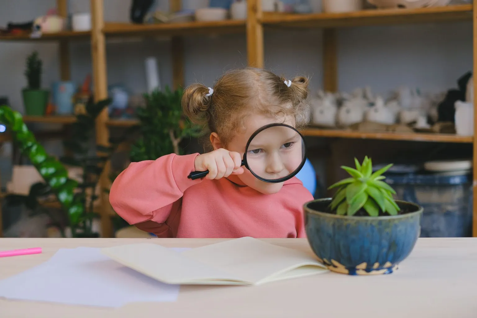Une petite fille observe une plante à travers une loupe, assise à une table.