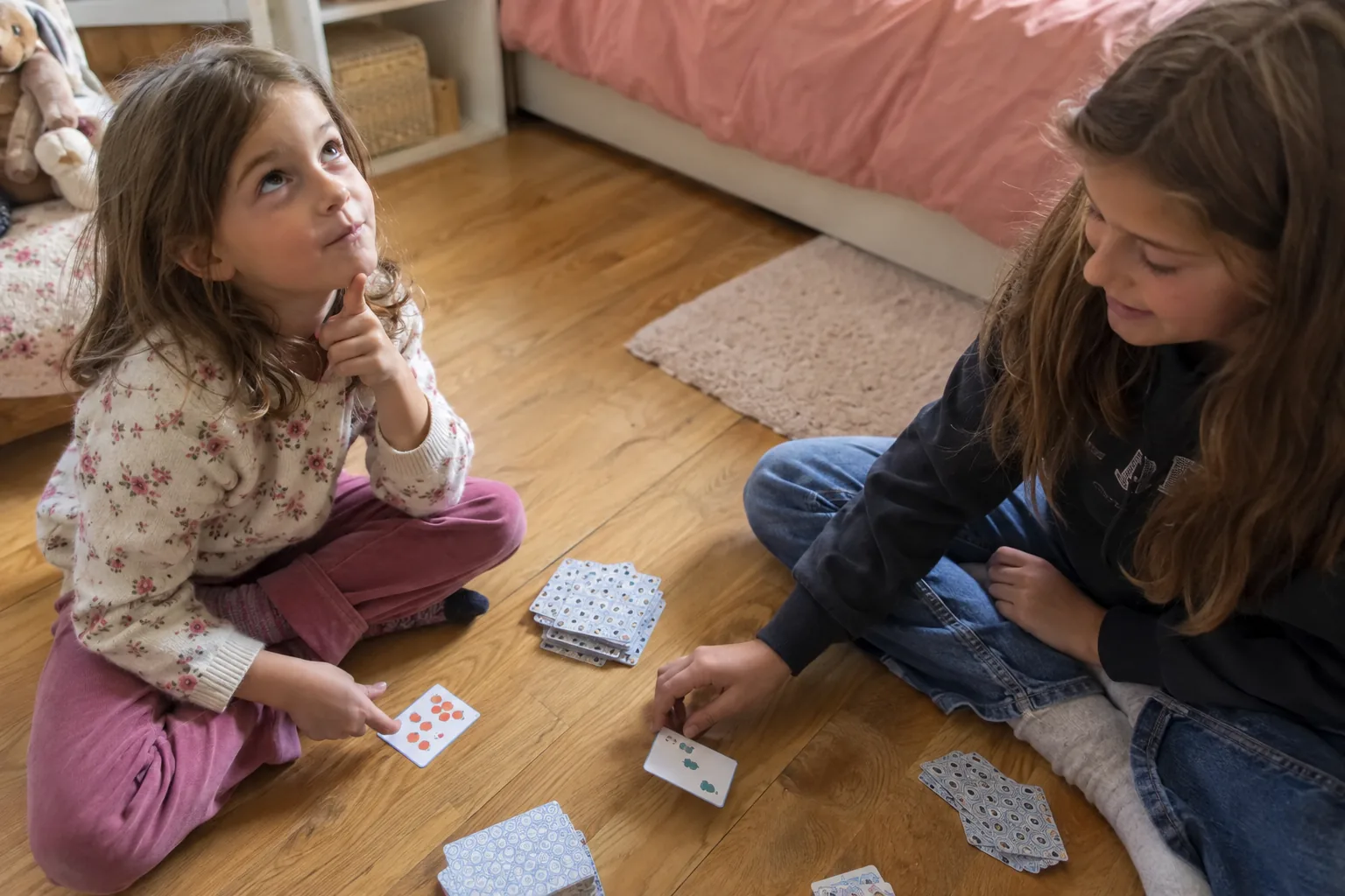 Deux sœurs jouent aux cartes sur le parquet, la plus jeune calcule de tête en regardant en l’air.